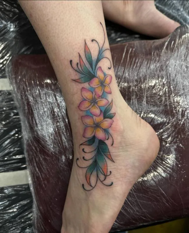 Illustrative color tattoo of clustered plumeria with peach, coral and teal leaves wrapped in fine black ornamental scrolls, on the ankle, by Julian (xwildxlovex) at Ohana Tattoo, Waikiki, Hawaii.
