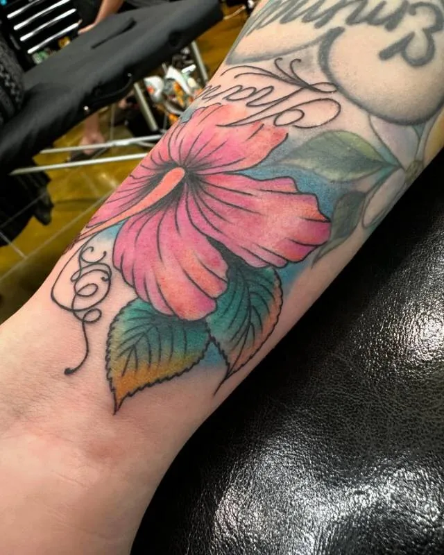 Illustrative color tattoo of a single oversized coral hibiscus with green leaves and watercolor sky behind it, on the inner forearm, by Julian (xwildxlovex) at Ohana Tattoo, Waikiki, Hawaii.