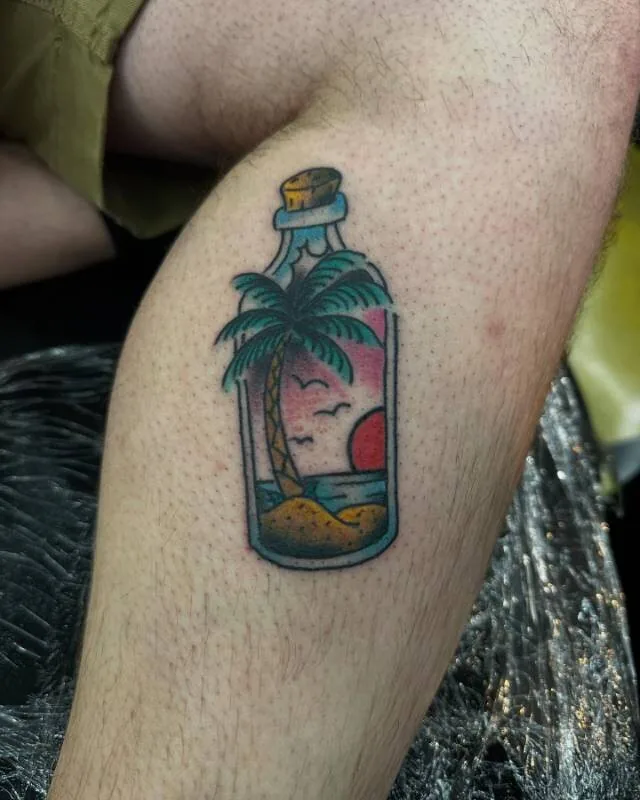 American Traditional flash tattoo of a tropical island scene inside a bottle with palm tree, red sunset and seagulls, on the lower leg, by Julian (xwildxlovex) at Ohana Tattoo, Waikiki, Hawaii.